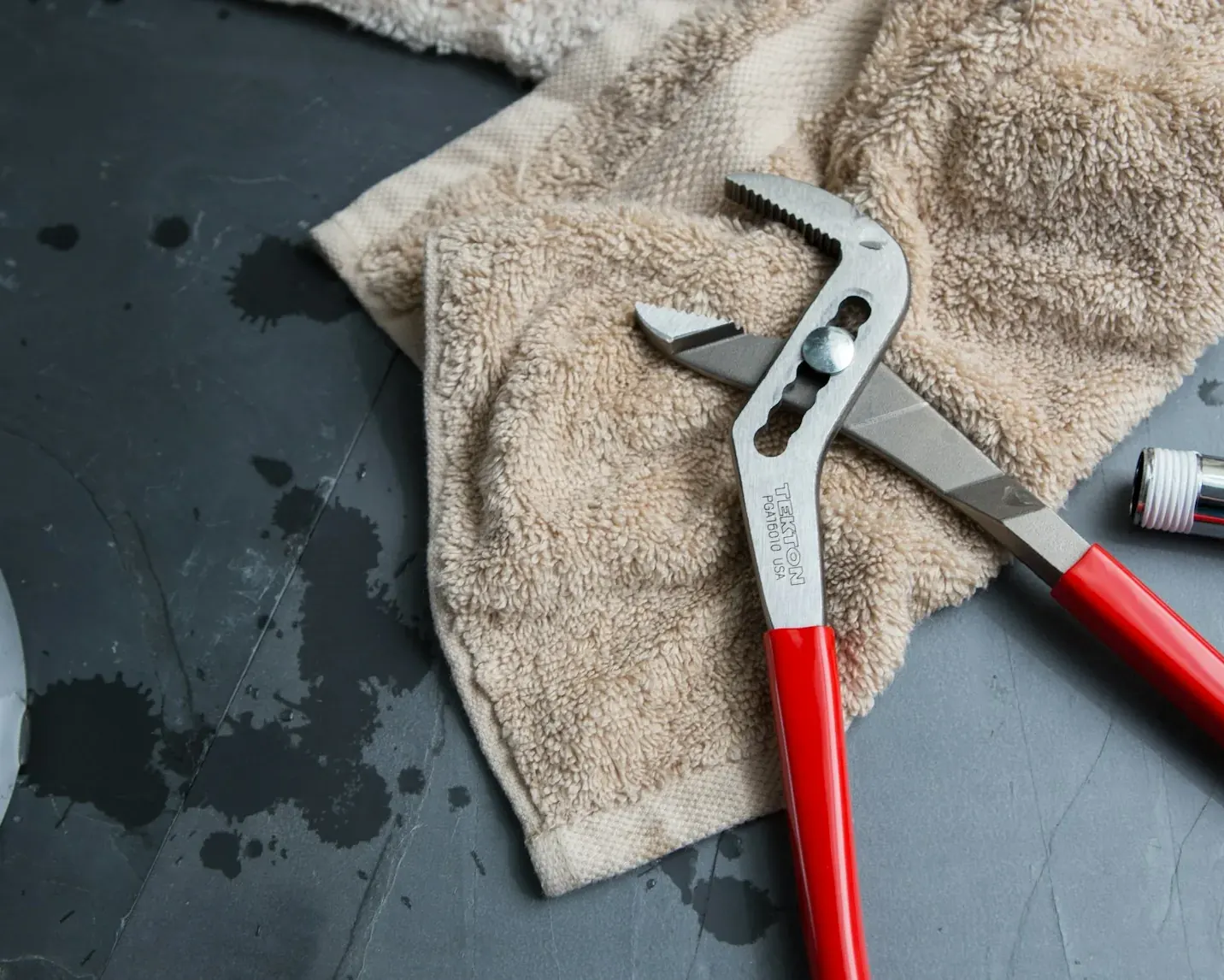 Red-handled pliers and a towel beside plumbing parts on a grey surface